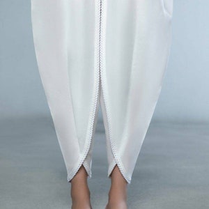 May include: White, flowing pants with a unique, draped design. The pants feature a decorative trim along the center seam and the bottom hem. The model is wearing embellished flats.