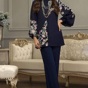 May include: A woman wearing a navy blue outfit with floral embroidery. She is wearing a long-sleeved top with a high neckline and matching wide-leg pants. The outfit is accented with a pearl necklace and sunglasses.