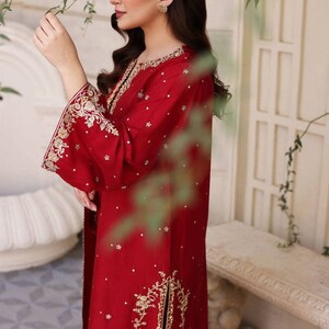 K&ouml;nnte beinhalten: Ein rotes, lang&auml;rmliges Kleid mit goldener Stickerei und Verzierungen. Das Kleid hat einen hohen Ausschnitt und einen langen Rock mit einem Schlitz an der Seite.