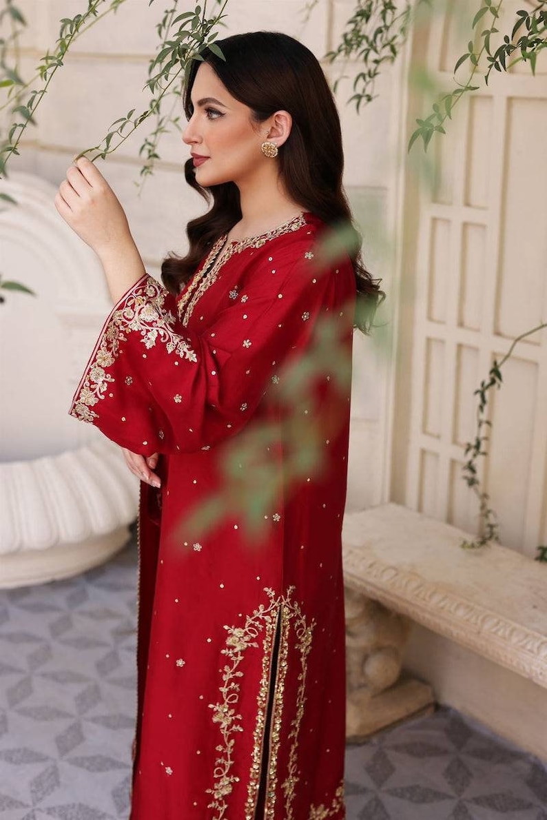 K&ouml;nnte beinhalten: Ein rotes, lang&auml;rmliges Kleid mit goldener Stickerei und Verzierungen. Das Kleid hat einen hohen Ausschnitt und einen langen Rock mit einem Schlitz an der Seite.
