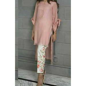 May include: A pink long-sleeved tunic with a high-low hemline and white trousers with floral embroidery. The tunic has a button fastening at the neckline and a tie detail on the sleeves.