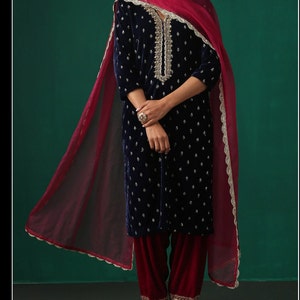 May include: A navy blue velvet kurta with silver embroidery and a matching maroon salwar. The kurta has a V-neckline and three-quarter sleeves. The salwar is loose-fitting and has a flared bottom. The outfit is paired with a maroon net dupatta with silver lace trim.