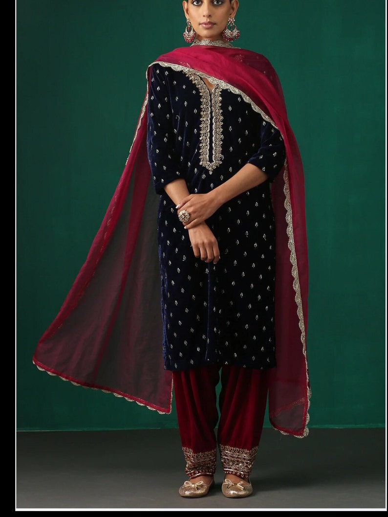 May include: A navy blue velvet kurta with silver embroidery and a matching maroon salwar. The kurta has a V-neckline and three-quarter sleeves. The salwar is loose-fitting and has a flared bottom. The outfit is paired with a maroon net dupatta with silver lace trim.