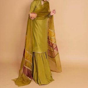 May include: A green salwar kameez with a sheer dupatta featuring gold and maroon accents. The dupatta has a geometric pattern and is draped over the shoulders.