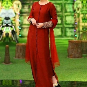 Puede incluir: Un vestido largo de color rojo con cierre de botones en la parte delantera y bordados dorados en las mangas. El vestido tiene una superposición de color rojo transparente y se lleva con un par de tacones blancos.