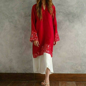 Puede incluir: Una túnica roja de manga larga con bordados blancos de paisley alrededor del cuello, el dobladillo y los puños. La túnica se lleva sobre una falda blanca.