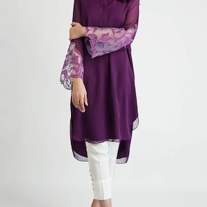May include: A long, purple tunic with a high neckline and long sleeves. The sleeves have a sheer, floral lace overlay. The tunic is paired with white pants.