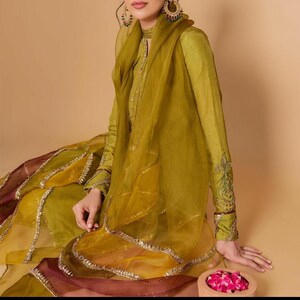 May include: A woman wearing a long, green, embroidered dress with a matching sheer scarf. The dress has a layered skirt with gold trim. The woman is sitting on a brown surface with a bowl of pink flowers in front of her.