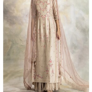 Pu&ograve; includere: Una kurta beige ricamente ricamata con un dupatta di velo rosa. La kurta ha un collo rotondo e maniche a tre quarti. Il dupatta ha un bordo in pizzo.