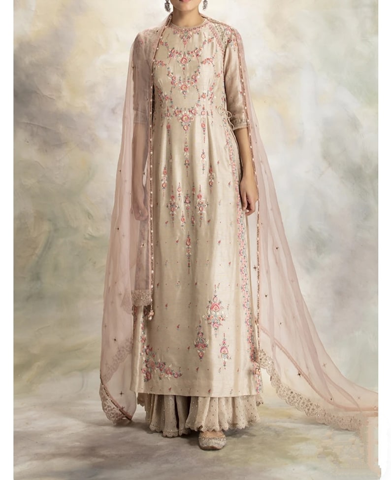 Pu&ograve; includere: Una kurta beige ricamente ricamata con un dupatta di velo rosa. La kurta ha un collo rotondo e maniche a tre quarti. Il dupatta ha un bordo in pizzo.