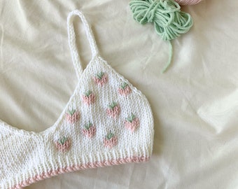 Puede incluir: Un top corto de punto blanco con fresas bordadas en rosa y verde. El top tiene un ribete rosa y tirantes finos.