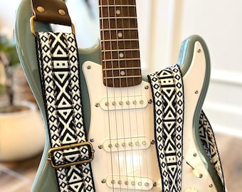 May include: A light blue electric guitar with a black and white patterned guitar strap. The guitar has a white pickguard and chrome hardware.