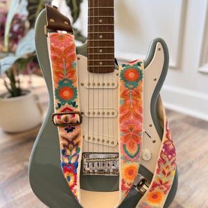May include: A sage green electric guitar featuring a white pickguard and a cream-colored guitar strap adorned with vibrant, multi-colored floral embroidery in shades of pink, orange, teal, and purple, secured with brown leather ends and metal hardware.