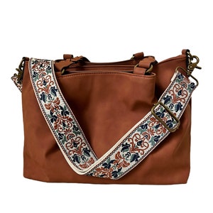 May include: A solid brown handbag featuring a wide, detachable shoulder strap with a cream-coloured background and a repeating floral pattern in shades of dark blue, rust orange, and green. The bag includes bronze-toned metal hardware and clips.
