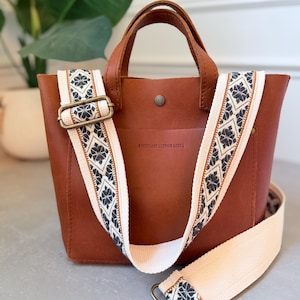 May include: A brown leather tote bag with two top handles and a detachable, adjustable woven shoulder strap. The strap features a cream, black, and orange geometric pattern. The bag is embossed with the text PORTLAND LEATHER GOODS.