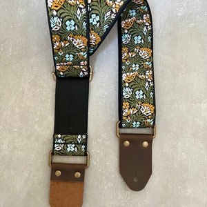 May include: A guitar strap with a floral pattern in shades of green, blue and yellow. The strap has brown leather ends with brass buckles.