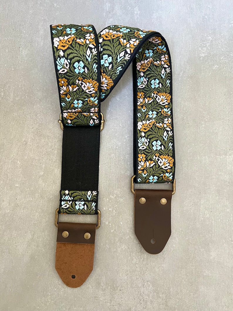 May include: A guitar strap with a floral pattern in shades of green, blue and yellow. The strap has brown leather ends with brass buckles.