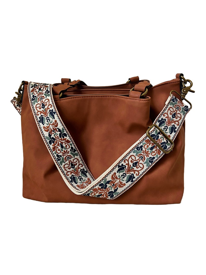 May include: A solid brown handbag featuring a wide, detachable shoulder strap with a cream-coloured background and a repeating floral pattern in shades of dark blue, rust orange, and green. The bag includes bronze-toned metal hardware and clips.