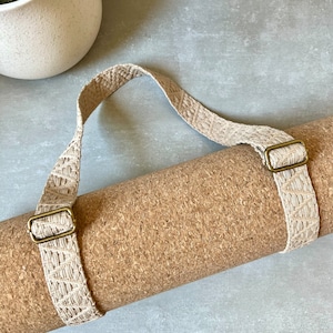 Op de afbeelding: Een opgerolde kurken yogamat met een beige geweven band en messing gespen. De band heeft een handvat om te dragen.