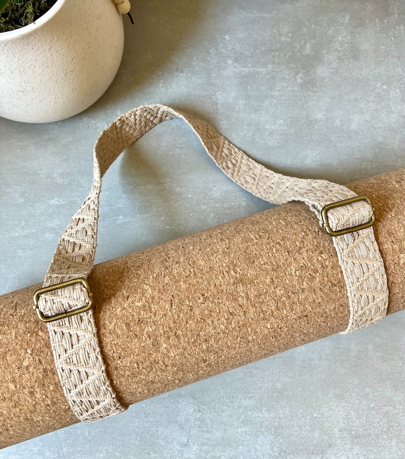 Op de afbeelding: Een opgerolde kurken yogamat met een beige geweven band en messing gespen. De band heeft een handvat om te dragen.