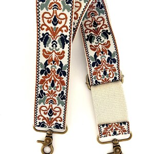 May include: A replacement bag strap featuring a cream-coloured base with an intricate woven pattern in burnt orange, navy blue, and sage green. The strap includes adjustable hardware and antique brass-toned metal swivel lobster claw clasps.