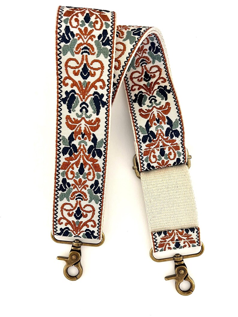 May include: A replacement bag strap featuring a cream-coloured base with an intricate woven pattern in burnt orange, navy blue, and sage green. The strap includes adjustable hardware and antique brass-toned metal swivel lobster claw clasps.
