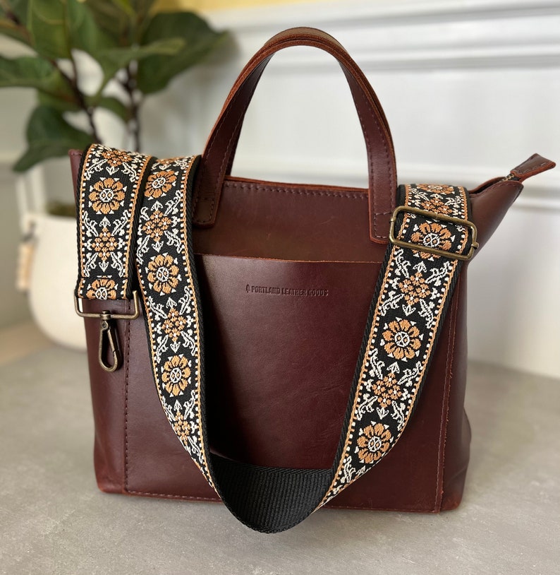 May include: A burgundy leather tote bag with a black and gold patterned strap. The bag has a small pocket on the front with the text "Portland Leather Goods".