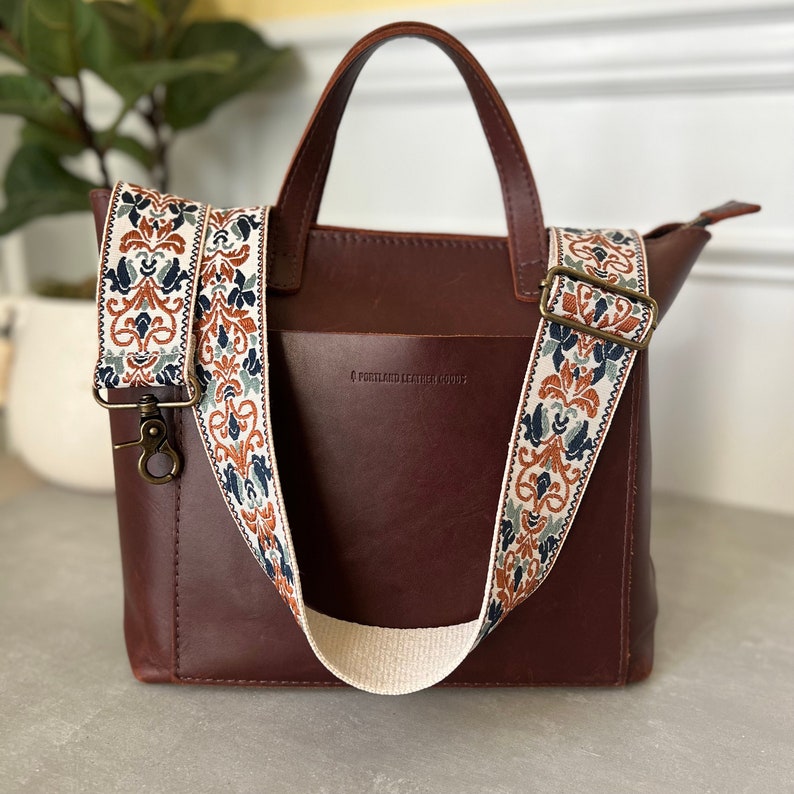 May include: A dark brown leather tote bag with two top handles and a detachable, wide woven shoulder strap. The strap features a cream, orange, and dark blue floral pattern. The bag has a front pocket and is embossed with Portland Leather Goods.