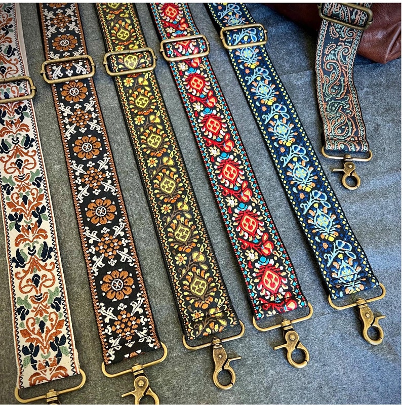 May include: Six colorful woven guitar straps with intricate patterns and gold metal clasps. The straps are arranged in a row, with the clasps facing the viewer.