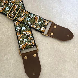 May include: A guitar strap with a floral pattern in shades of green, blue, and yellow. The strap has brown leather ends with brass buckles.