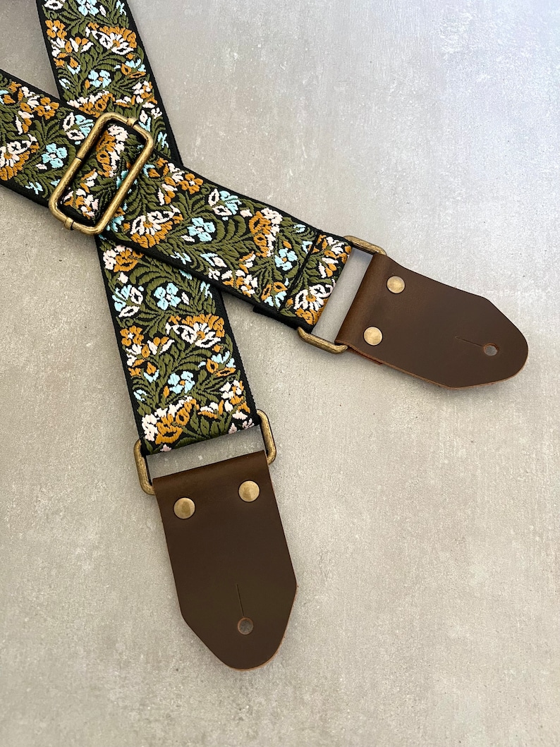 May include: A guitar strap with a floral pattern in shades of green, blue, and yellow. The strap has brown leather ends with brass buckles.