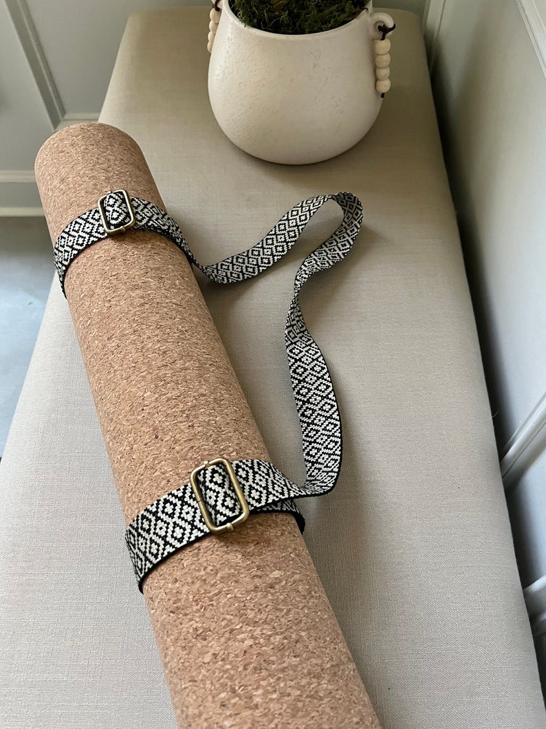 Op de afbeelding: Een bruine kurk yogamat met een zwart-wit gepatroneerde band met gouden gespen.