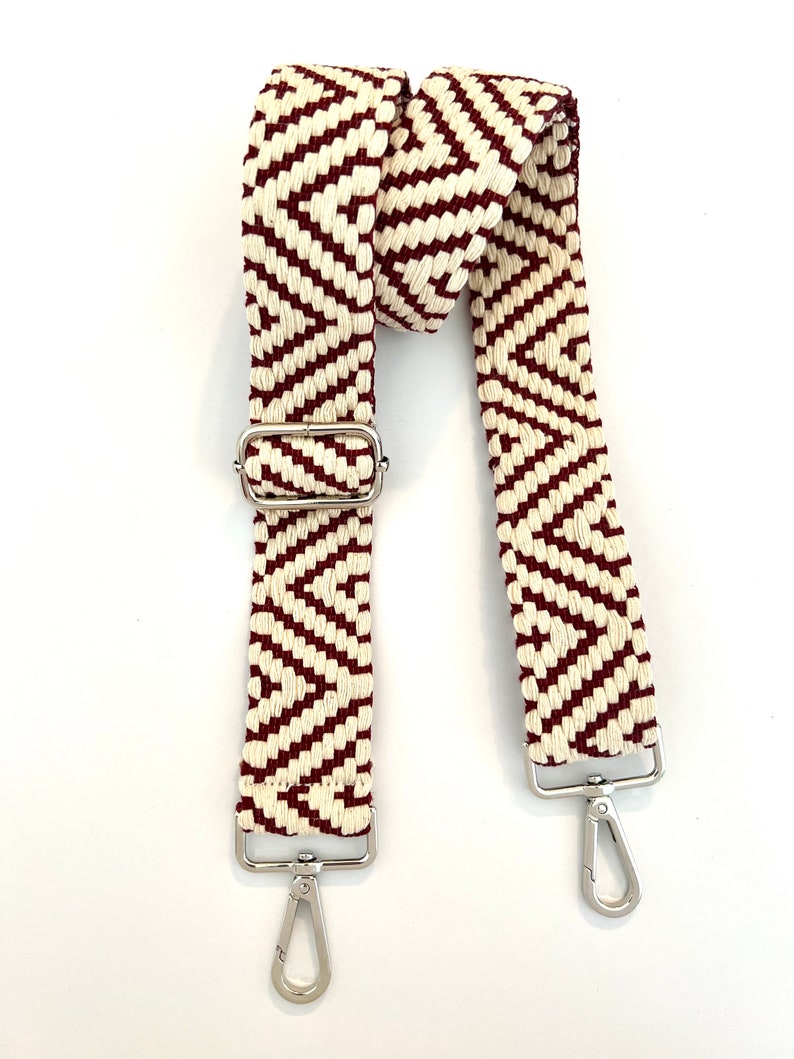 Op de afbeelding: Een geweven band met rood en wit chevron-patroon met zilveren metalen hardware, waaronder een gesp en twee draaibare clips.