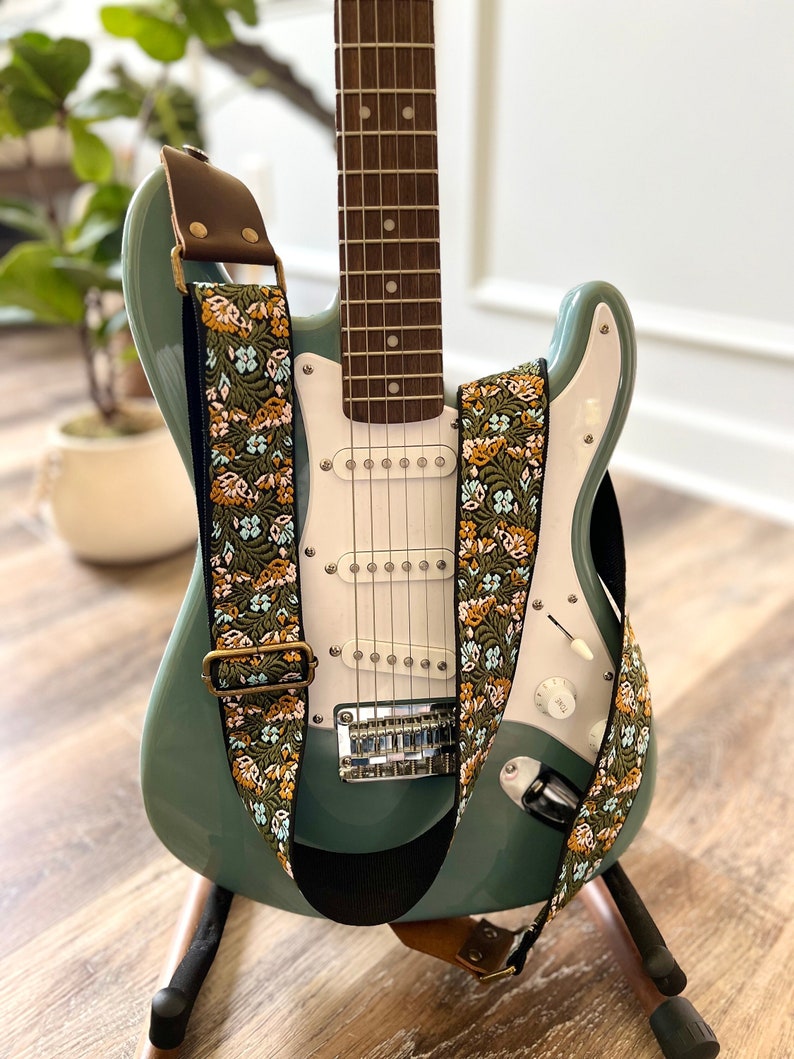May include: A teal electric guitar with a floral patterned guitar strap. The strap is brown, green, and orange with a gold buckle.