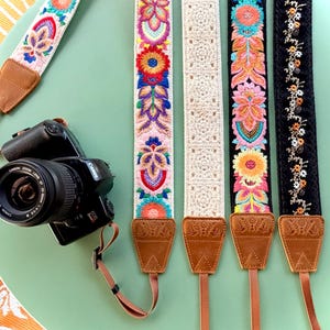 May include: Five camera straps with brown leather ends and various designs including colourful floral embroidery on white and black fabric, and a cream-coloured crocheted pattern. A black Canon camera is attached to one strap.