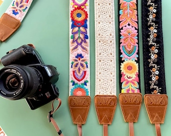 May include: Five camera straps with brown leather ends and various designs including colorful floral embroidery on white and black fabric, and a cream-colored crocheted pattern. A black Canon camera is attached to one strap.
