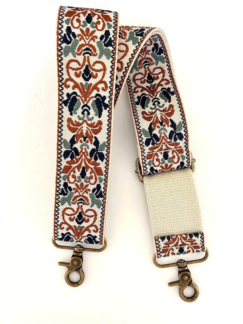 May include: A white camera strap with a brown, blue, and green floral pattern. The strap has two gold metal clasps.