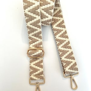 Op de afbeelding: Een geweven tasriem met beige en wit patroon en gouden metalen hardware. De riem heeft twee verstelbare gespen en twee gouden metalen sluitingen.