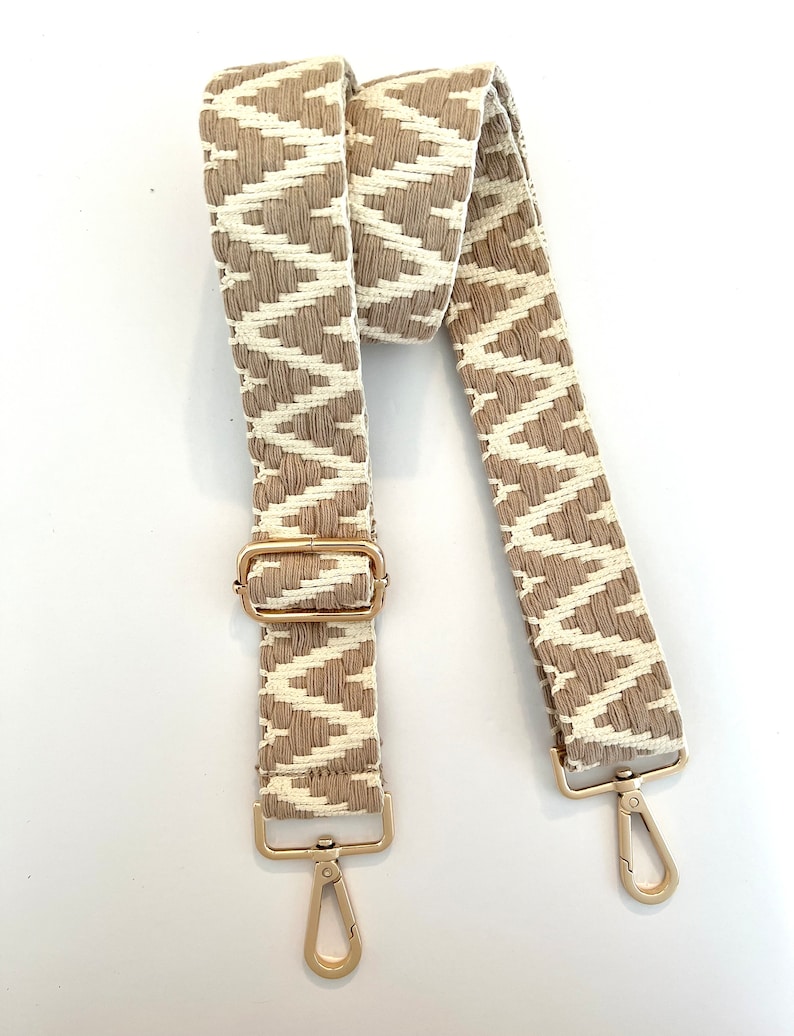 Op de afbeelding: Een geweven tasriem met beige en wit patroon en gouden metalen hardware. De riem heeft twee verstelbare gespen en twee gouden metalen sluitingen.