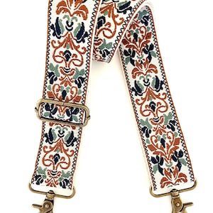 May include: A wide, adjustable bag strap featuring a cream-coloured base with an intricate woven pattern in shades of burnt orange, navy blue, and sage green. The strap includes antique brass-toned metal hardware, including a slider and two swivel clips.