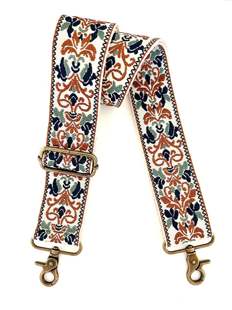 May include: A wide, adjustable bag strap featuring a cream-coloured base with an intricate woven pattern in shades of burnt orange, navy blue, and sage green. The strap includes antique brass-toned metal hardware, including a slider and two swivel clips.