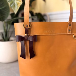 Könnte beinhalten: Eine hellbraune Ledertasche mit weißen Nähten und zwei Schulterriemen. Ein dunkelbrauner Lederanhänger in Schleifenform ist an einem Messingring an der Vorderseite befestigt. Der Schriftzug PORTLAND LEATHER ist oben auf der Tasche eingeprägt.