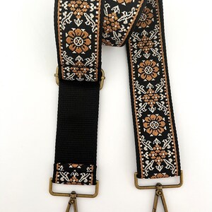 May include: Black and brown woven strap with gold-toned metal clasps and hooks. The strap is designed with a floral pattern.
