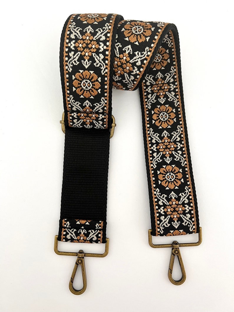 May include: Black and brown woven strap with gold-toned metal clasps and hooks. The strap is designed with a floral pattern.