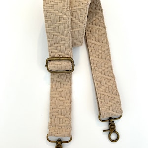 Op de afbeelding: Een beige geweven band met twee gouden metalen gespen en een gouden metalen gesp.