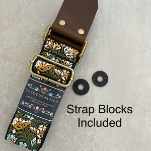 May include: A black and floral guitar strap with a brown leather end and two black strap blocks. The strap is handmade in the USA with genuine leather. The text "TF CO GUITAR STRAP" is printed on a white label attached to the strap. "Strap Blocks Included" is printed in black text on a white background.