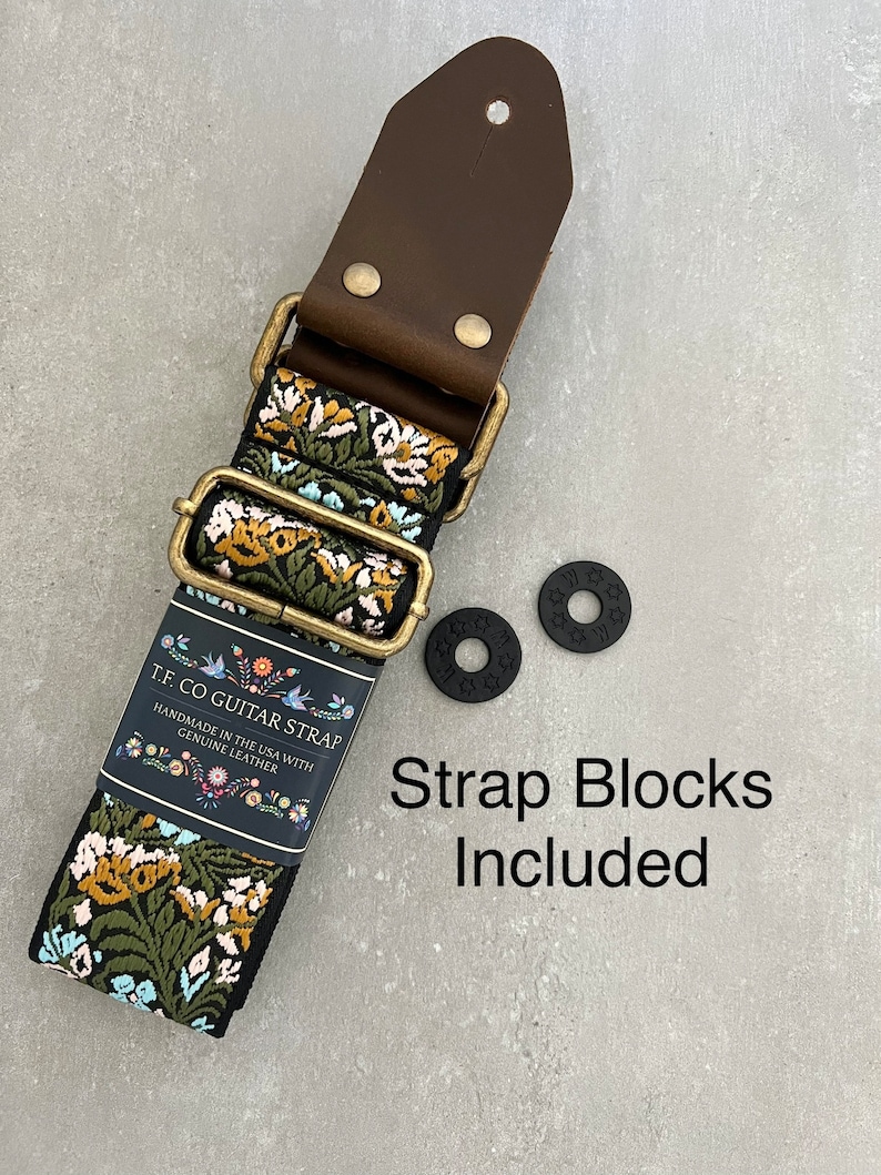 May include: A black and floral guitar strap with a brown leather end and two black strap blocks. The strap is handmade in the USA with genuine leather. The text "TF CO GUITAR STRAP" is printed on a white label attached to the strap. "Strap Blocks Included" is printed in black text on a white background.
