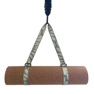 Op de afbeelding: Een bruine yogamat met een grijze en witte gepatenteerde band die aan een zwarte veer-vormige haak hangt.
