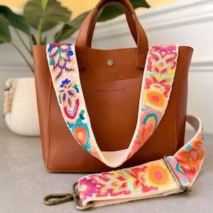 May include: A brown leather tote bag with two top handles and a detachable, wide shoulder strap featuring vibrant, multi-coloured floral embroidery. The bag has a central snap closure and the text Portland Leather Goods embossed on the front.