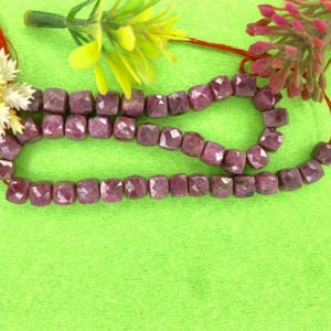 Può includere: Una collana di perle di rubino rosa, ogni perla è sfaccettata e di forma quadrata. Le perle sono infilate su un filo rosso e sono esposte su una superficie verde.